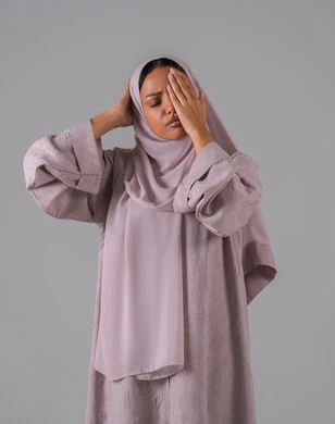 الاحساس بالشقيقية والصداع النصفي, الشعور بالإرهاق وفقدان التركيز, الاجهاد والضغط النفسي,  بورتريه لامرأه عربيه خليجيه سعوديه ترتدي العباءة والحجاب تضع يديها على رأسها وتغمض عينيها بإيماءات ألم الرأس الشديد, خلفية رمادية