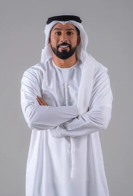الوقوف باستقامة وايدي متقاطعة، مفهوم الرجولة والثقة بالنفس, بورتريه لرجل عربي خليجي إماراتي يرتدي الكندورة والغترة البيضاء ينظر بتعابير السرور، الأناقة والاهتمام بالمظهر الخارجي، خلفية رماديه.