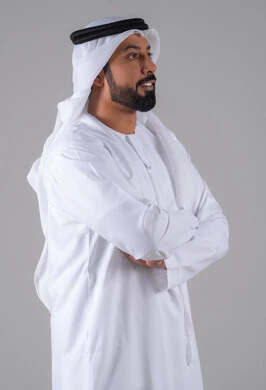 بورتريه لرجل عربي خليجي إماراتي يرتدي الكندورة والغترة البيضاء ينظر بتعابير السرور، الوقوف باستقامة وايدي متقاطعة، مفهوم الرجولة والثقة بالنفس,الأناقة والاهتمام بالمظهر الخارجي، خلفية رماديه.