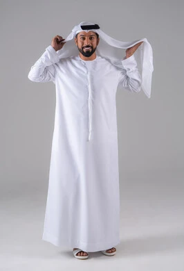 مفهوم الرجولة والثقة بالنفس, بورتريه لرجل عربي خليجي إماراتي يرتدي الكندورة ويعدل من وضعية الغترة البيضاء ينظر بتعابير السرور، الأناقة والاهتمام بالمظهر الخارجي، خلفية رماديه.