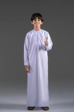 نظرات السرور والشعور بالرضى, بورتريه لولد عربي خليجي سعودي يرتدي الثوب التقليدي الابيض  يرفع إبهامه للأعلى بعلامة الجودة, الوقوف باستقامة والنظر إلى الكاميرا بإيماءات التفاعل والموافقة, صورة الجسم بالطول الكامل, خلفية رمادية