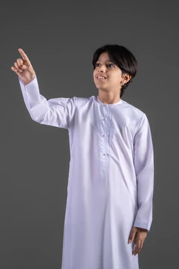 تحديد موقع والاشارة اليه, الاشارة الى مكان ما بواسطة اصبع السبابة, بورتريه  لولد عربي خليجي سعودي يرتدي الثوب الابيض التقليدي يشير باصبعه لشيء ما بايماءات السعادة, اشارات وحركات معبرة،الاناقة والاهتمام بالمظهر الخارجي، الجسم بالطول الكامل, خلفية رمادي