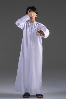 إغلاق العيون باستخدم اليدين، عدم وضوح الرؤية بشكل جيد، الم في العيون والرأس ، التركيز على شيء ما، بورتريه لولدعربي خليجي سعودي يرتدي الثوب التقليدي  يضع يده على عينيه بشكل  بإيماءات عدم العثور على شيء، الجسم بالطول الكامل، خلفية رمادي