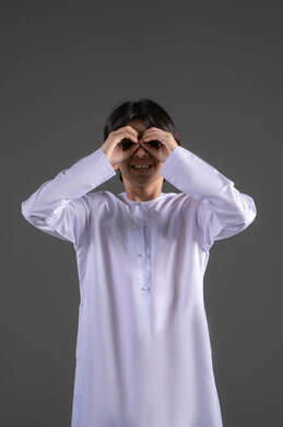 محاولة تقريب الأشياء البعيدة، التخطيط للأهداف والطموحات المستقبلية، حب الاكتشاف والمعرفة والمغامرة، التركيز على شيء ما، بورتريه لولدعربي خليجي سعودي يرتدي الثوب التقليدي  يضع يده على عينيه بشكل عدسة بإيماءات البحث، الجسم بالطول الكامل، خلفية رمادي