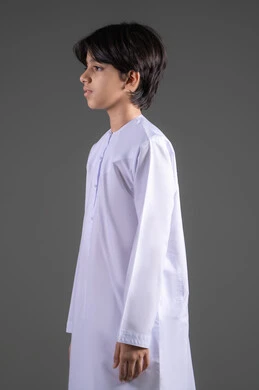 Expressions of handsome and manliness since childhood, getting ready and preparing to go outside, taking care of the external appearance, looking at the camera with gestures of happiness and joy, a side portrait of an Arab Gulf Saudi boy wearing the traditional white thobe, full body, gray background.