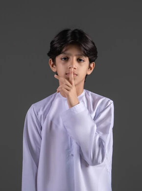 الهدوء والسكون وعدم الإزعاج، طلب السكوت والصمت، بورتريه  لفتى عربي خليجي سعودي يرتدي الثوب التقليدي يضع أصبع السبابة على فمه، عدم التدخل وخفض الصوت والتهديد, حركة أصبع وإشارات، التوقف عن الكلام وخفض الصوت، خلفية رماديه