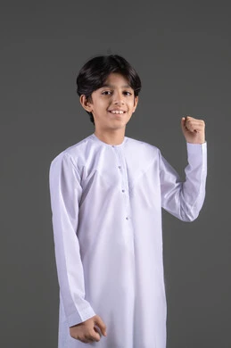 Increasing the muscles' endurance, victory, winning, and success, the concept of achievement and goal realization, a portrait of a smiling Arab Gulf Saudi boy wearing a traditional white thobe raising his fist in the air with gestures of joy and happiness, encouragement with expressions of enthusiasm, feeling of happiness and strength, gray background.