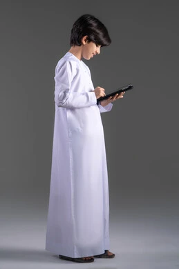 قضاء وقت ممتع بالتسلية على الإنترنت، التعلم عن بعد،  الاستمتاع في متابعه البرامج الترفيهيه المفضلة، بورتريه لولد عربي خليجي سعودي مبتسم يرتدي الثوب التقليدي يحمل بيده جهاز التابليت، استخدام التقنيات الحديثة في تزويد الأجيال بالمعلومات المفيدة، خلفية رمادية