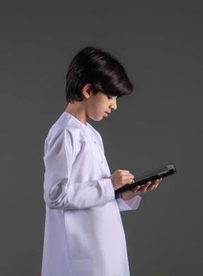 التعلم عن بعد،  الاستمتاع في متابعه البرامج الترفيهيه المفضلة، بورتريه لولد عربي خليجي سعودي مبتسم يرتدي الثوب التقليدي يحمل بيده جهاز التابليت، استخدام التقنيات الحديثة في تزويد الأجيال بالمعلومات المفيدة، قضاء وقت ممتع بالتسلية على الإنترنت، خلفية رمادية