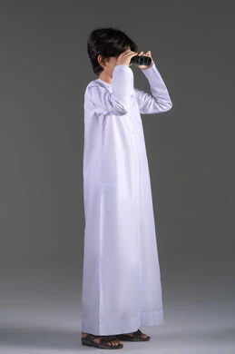 توضيح مجال الرؤية باستخدام المنظار، رؤية الاشياء البعيدة عبر المنظار، التخطيط المستقبلي للأهداف والطموحات، بورتريه جانبي لولد عربي خليجي سعودي مبتسم يرتدي الثوب التقليدي يحمل بيده المنظار ثنائي العينين، حب الاستكشاف والمغامرة، الجسم بالطول الكامل، خلفية رمادية