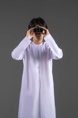 تعابير التركيز والتفاعل، رؤية الاشياء البعيدة عبر المنظار، التخطيط المستقبلي للأهداف والطموحات، بورتريه لولد عربي خليجي سعودي مبتسم يرتدي الثوب التقليدي يحمل بيده المنظار ثنائي العينين، حب الاستكشاف والمغامرة، الجسم بالطول الكامل، خلفية رمادية