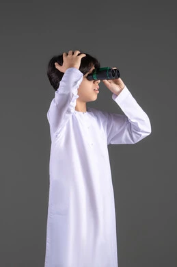 رؤية الاشياء البعيدة عبر المنظار، حب المغامرة والاستكشاف،  بورتريه لولد عربي خليجي اماراتي يرتدي الكندورة  ينظر من خلال المنظور ثنائي العينين (التلسكوب) بإيماءات الفرح والسرور، التخطيط المستقبلي للأهداف والطموحات، خلفية رمادية