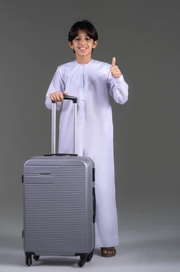 Thumbs up with gestures of admiration, a portrait of an Emirati Gulf Arab boy wearing a kandura holding a travel bag, enjoying a fun recreational trip, wellness and travel trips, gray background.