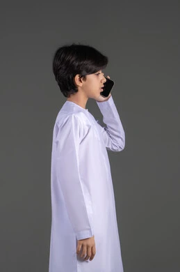 التواصل والدردشة مع الأصدقاء والاقارب، بورتريه لشاب عربي خليجي اماراتي  يرتدي الثوب التقليدي يقوم بإجراء مكالمة هاتفية عبر الجوال، قضاء وقت ممتع بتبادل الأحاديث، خلفية رمادية