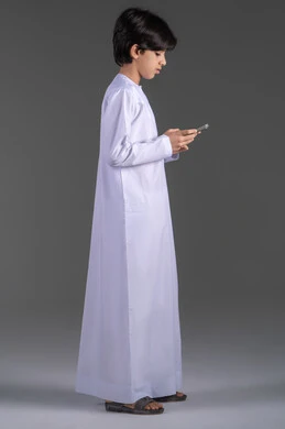 Spending enjoyable time using the phone, communicating and chatting with friends and relatives, a portrait of a young Emirati Arab man wearing traditional attire using the phone for something, with a gray background.