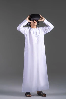 Simulation of the imaginary world, enjoyment and entertainment through viewing with virtual reality glasses, a portrait of an Emirati Gulf Arab boy wearing a kandura looking through 3D glasses (VR) with gestures of joy, using a modern and advanced technical device, gray background.