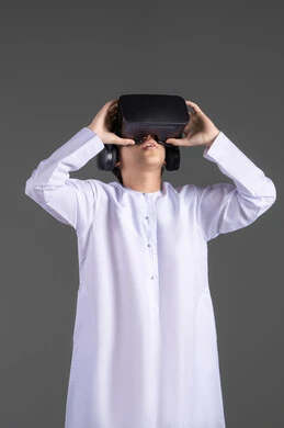 The use of a modern and advanced technical device, simulating the imaginary world, enjoyment and entertainment through viewing via virtual reality glasses, a portrait of an Emirati Gulf Arab boy wearing a kandura looking through 3D glasses (VR) with gestures of joy, gray background.