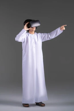 Simulation of the imaginary world, enjoyment and entertainment through viewing with virtual reality glasses, a portrait of an Emirati Gulf Arab boy wearing a kandura looking through 3D glasses (VR) with gestures of joy, using a modern and advanced technical device, gray background.