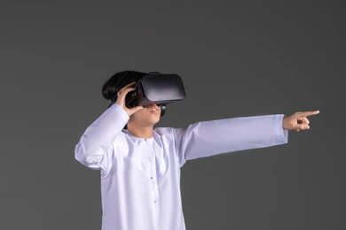 The use of a modern and advanced technical device, simulating the imaginary world, enjoyment and entertainment through viewing via virtual reality glasses, a portrait of an Emirati Gulf Arab boy wearing a kandura looking through 3D glasses (VR) with gestures of joy, gray background.