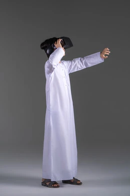 Simulation of the imaginary world, enjoyment and entertainment through viewing with virtual reality glasses, a portrait of an Emirati Gulf Arab boy wearing a kandura looking through 3D glasses (VR) with gestures of joy, using a modern and advanced technical device, gray background.