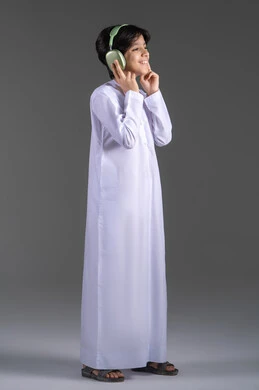 التسلية والترفيه بإستخدام تقنية الهيدفون، سماع الموسيقى والصوتيات، ارتداء سماعات الرأس الصوتية، بورتريه لولد عربي خليجي إماراتي يرتدي الكندورة الإماراتية يقوم بالاستماع للأغاني بايماءات السرور والسعاة، خلفية رمادية