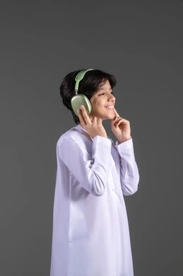 سماع الموسيقى والصوتيات،التسلية والترفيه بإستخدام تقنية الهيدفون،  ارتداء سماعات الرأس الصوتية، بورتريه لولد عربي خليجي إماراتي يرتدي الكندورة الإماراتية يقوم بالاستماع للأغاني بايماءات السرور والسعاة، خلفية رمادية