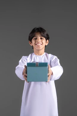 Gift giving during holidays and occasions, expressing warmth and love, a portrait of a smiling Emirati Gulf Arab boy wearing traditional attire holding a blue gift box, gray background.