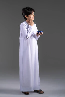 روتين العناية بالبشرة،العناية والإهتمام بنضارة وتحسين البشرة، بورتريه لولد عربي خليجي اماراتي يرتدي الثوب التقليدي , يضع كريم الترطيب على  الوجه، ترطيب وتفتيح البشرة،صورة الجسم كامل, خلفيه رمادية