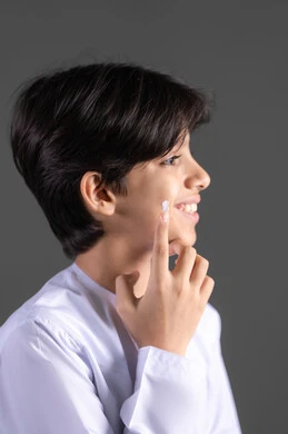 Caring for and improving skin freshness, a close-up portrait of an Emirati Gulf Arab boy wearing traditional attire, applying moisturizer to his face, skincare routine, moisturizing and brightening the skin, gray background.
