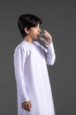Detoxifying the body, maintaining and treating kidney health, a portrait of an Emirati Gulf Arab boy wearing a kandura drinking a glass of water, the importance of drinking pure water for achieving radiant skin, preserving the integrity of the immune system, the full-length body, gray background.