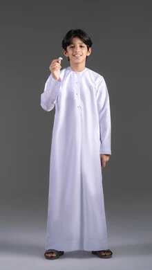 The concept of using mobile device accessories, a portrait of an Emirati Gulf Arab boy wearing traditional attire, looking at the camera with gestures of joy and holding a modern mobile Bluetooth headset in his hand, full-body, gray background. The concept of using mobile device accessories, a portrait of an Emirati Gulf Arab boy wearing traditional attire, looking at the camera with gestures of joy and holding a modern mobile Bluetooth headset in his hand, full-body, gray background.