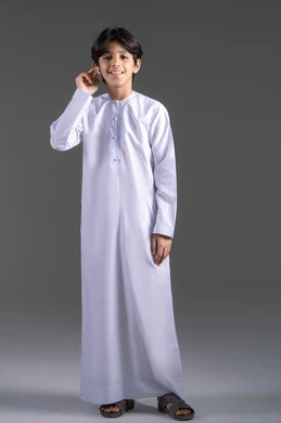 مفهوم إستخدام إكسسوارات الجهاز المحمول، بورتريه لولد  عربي خليجي اماراتي يرتدي الزي التقليدي، ينظر الى الكاميرا بايماءات السرور ويحمل بيده سماعة بلوتوث جوال حديثه،جسم بالطول الكامل،  خلفيه رماديه