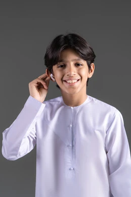 بورتريه لولد  عربي خليجي اماراتي يرتدي الزي التقليدي، ينظر الى الكاميرا بايماءات السرور ويحمل بيده سماعة بلوتوث جوال حديثه، مفهوم إستخدام إكسسوارات الجهاز المحمول، خلفيه رماديه