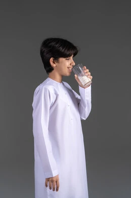 مفهوم العناية والاهتمام بالصحة, أهمية شرب الحليب عند الاولاد, إتباع نظام غذائي صحي, بورتريه من الجانب لولد عربي خليجي اماراتيي مبتسم يرتدي الكندورة يقوم بشرب كوب حليب , خلفية رمادية