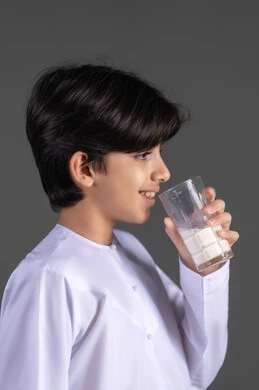 أهمية شرب الحليب عند الاولاد, إتباع نظام غذائي صحي, بورتريه مقرب من الجانب لولد عربي خليجي اماراتيي مبتسم يرتدي الكندورة يقوم بشرب كوب حليب , مفهوم العناية والاهتمام بالصحة, خلفية رمادية