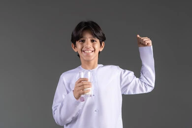 أهمية شرب الحليب عند الأطفال, النظر الى الكاميرا بإيماءات السرور، إتباع نظام غذائي صحي, بورتريه مقرب لولد عربي خليجي اماراتيي مبتسم يرتدي الكندورة يحمل بيده كوب حليب , مفهوم العناية والاهتمام بالصحة, خلفية رمادية