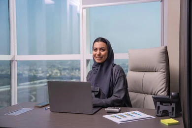 مهنة ووظيفة نسائية، ادارة الأعمال وانجاز المهام المطلوبة، إمرأة عربية خليجية اماراتية محجبة ترتدي عباءة تجلس على المكتب تستخدم الحاسوب المحمول، استخدام جهاز تقني حديث ومتطور، ايماءات التركيز والاندماج