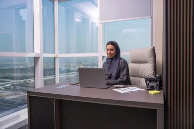 ادارة الأعمال وانجاز المهام المطلوبة، مهنة ووظيفة نسائية، إمرأة عربية خليجية اماراتية محجبة ترتدي عباءة تجلس على المكتب تستخدم الحاسوب المحمول، استخدام جهاز تقني حديث ومتطور، ايماءات التركيز والاندماج