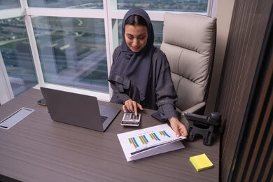 Completing calculations using a calculator, the analytics and statistical graphics department, an Arab Gulf Emirati woman wearing an abaya and hijab working on managing accounts, using a laptop, office professions and jobs, the stock market and financial investment.