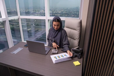 Using the calculator to complete numerical operations, the analytics and statistical graphics department, an Arab Gulf Emirati woman wearing an abaya and hijab working on account management, using a laptop, office professions and jobs, the stock market and financial investment.