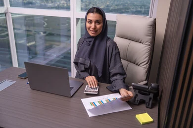 إجراء الحسابات بواسطة الآلة الحاسبة، قسم التحليلات والإحصائيات البيانية،امراه عربية خليجية اماراتية ترتدي العباءة و الحجاب تعمل على إدارة الحسابات، إستخدام جهاز الحاسوب المحمول، مهن ووظائف مكتبية، البورصة والإستثمار المالي