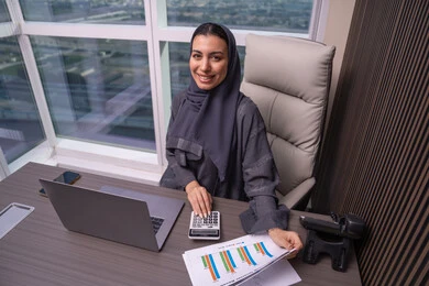 Performing calculations using a calculator, the analytics and statistical graphics department, an Arab Gulf Emirati woman wearing an abaya and hijab working on account management, using a laptop, office professions and jobs, the stock market and financial investment.