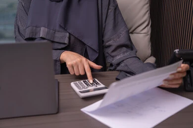 أداء العمليات الحسابية عبر الآلة الحاسبة، مهن ووظائف مكتبية، قسم التحليلات والإحصائيات البيانية، صورة مقربة ليد امراه عربية خليجية اماراتية ترتدي العباءة تعمل على إدارة الحسابات، إستخدام جهاز الحاسوب المحمول، البورصة والإستثمار المالي