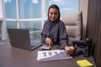 إستخدام الآلة الحاسبة لإجراء العمليات الحسابية، إستخدام جهاز الحاسوب المحمول، قسم التحليلات والإحصائيات البيانية،امراه عربية خليجية اماراتية ترتدي العباءة و الحجاب تعمل على إدارة الحسابات، مهن ووظائف مكتبية، البورصة والإستثمار المالي