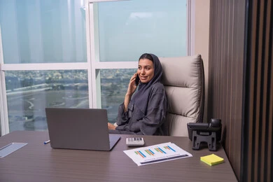 Completing work via mobile, enjoying exchanging conversations and news, an Arab Gulf Emirati woman wearing an abaya and hijab sits at the desk making a phone call using her mobile phone, a comfortable work environment in Emirati companies.