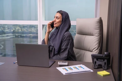 Remote mobile work, enjoying exchanging conversations and news, an Arab Gulf Emirati woman wearing an abaya and hijab sitting at a desk making a phone call using a mobile phone, a comfortable work environment in Emirati companies.