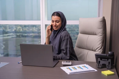 Following up on work remotely via phone, enjoying exchanging conversations and news, an Arab Gulf Emirati woman wearing an abaya and hijab sits at the desk making a phone call using a mobile phone, a comfortable work environment in Emirati companies.