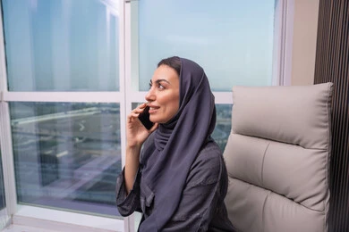 تنفيذ المهام الوظيفية من خلال الجوال، الاستمتاع بتبادل الأحاديث و الأخبار، امراه عربيه خليجيه اماراتية ترتدي العباءة و الحجاب تجلس على المكتب تقوم بإجراء مكالمة هاتفيه باستخدام الهاتف المحمول, بيئة عمل مريحة في الشركات الاماراتية