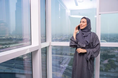 بيئة عمل مريحة في الشركات الاماراتية، الاستمتاع بتبادل الأحاديث و الأخبار، العمل عن بعد عبر الجوال، امراه عربيه خليجيه اماراتية ترتدي العباءة و الحجاب تقف بجانب النافذة تقوم بإجراء مكالمة هاتفيه باستخدام الهاتف المحمول