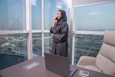 Enjoying exchanging conversations and news, working remotely via mobile, an Arab Gulf Emirati woman wearing an abaya and hijab stands by the window making a phone call using her mobile phone, a comfortable work environment in Emirati companies.